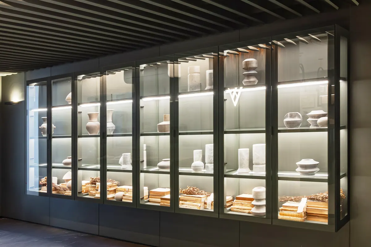 Cristaleira longa com portas de vidro, prateleiras iluminadas e objetos decorativos em ambiente escuro