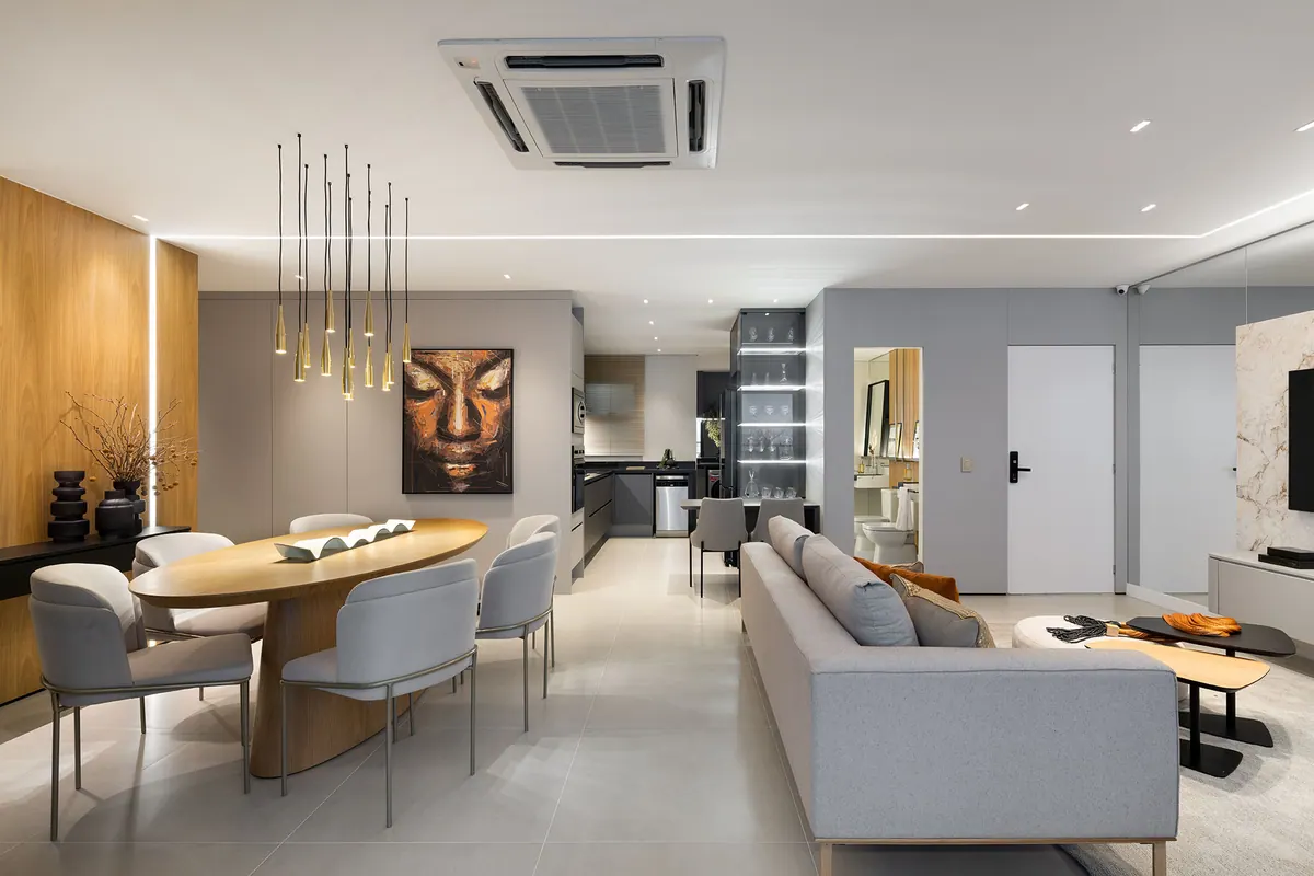 Sala integrada com painel em tom amadeirado, mesa de jantar oval e armários da cozinha ao fundo