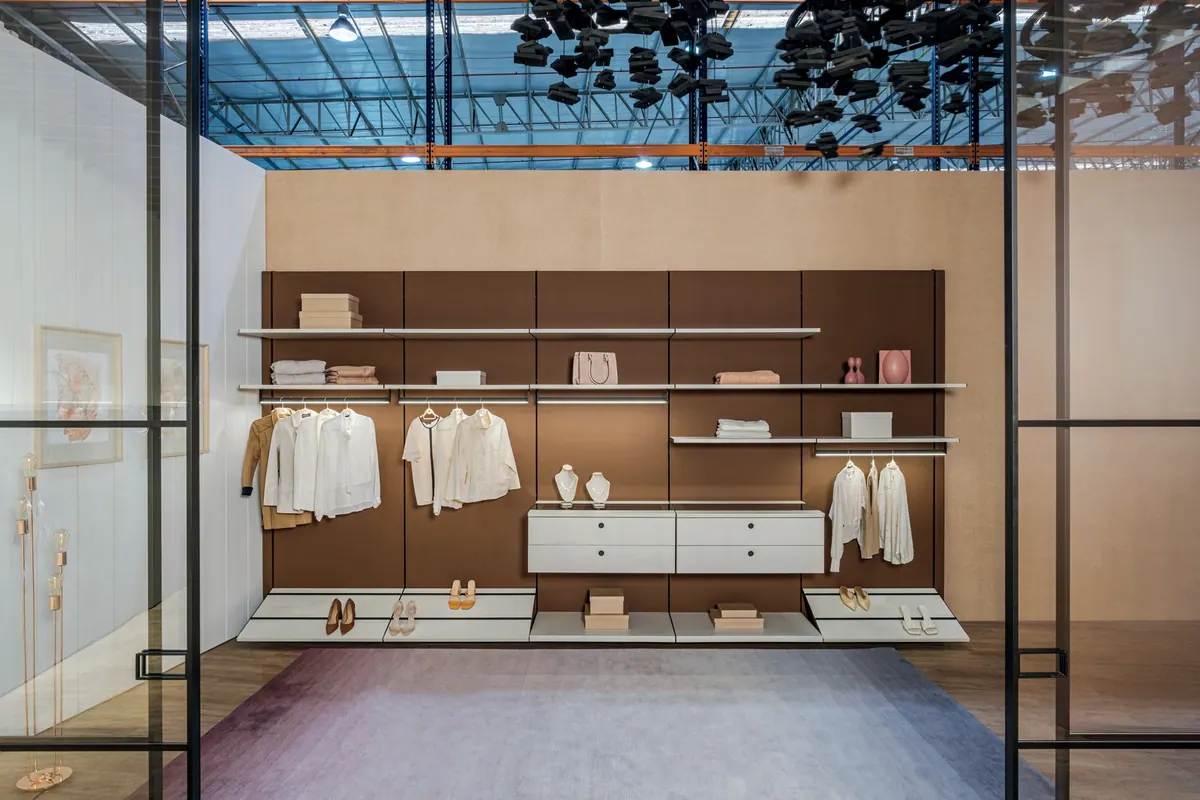 Closet aberto com painel marrom, prateleiras, cabideiros e gaveteiro, visto de frente por divisória de vidro