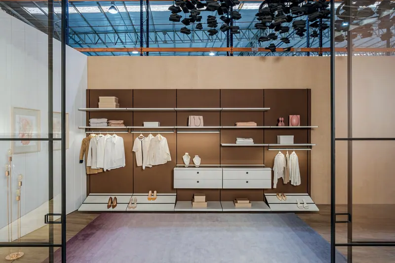 Closet aberto com painel marrom, prateleiras, cabideiros e gaveteiro, visto de frente por divisória de vidro