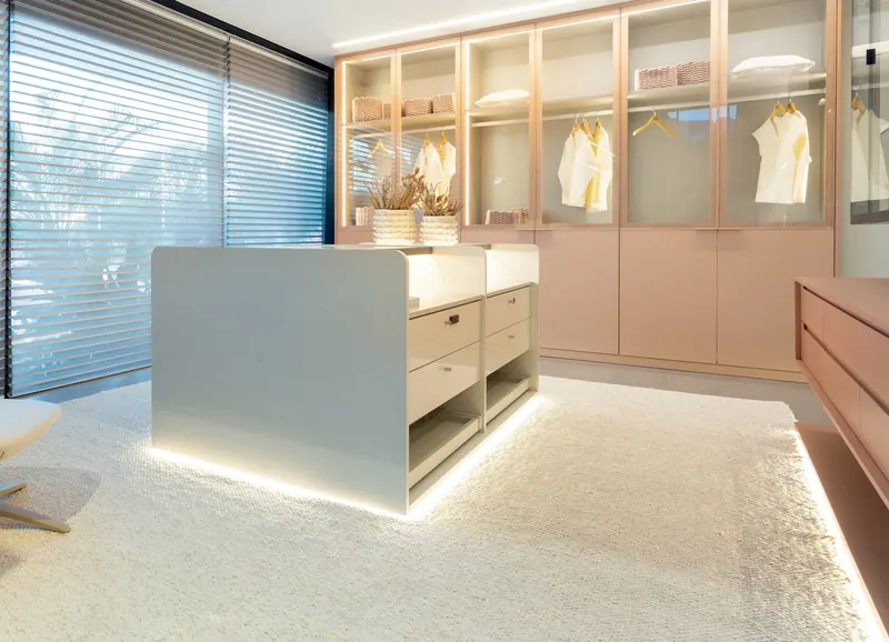 Closet com armários rosados de portas de vidro, ilha central com gavetas e led no rodapé