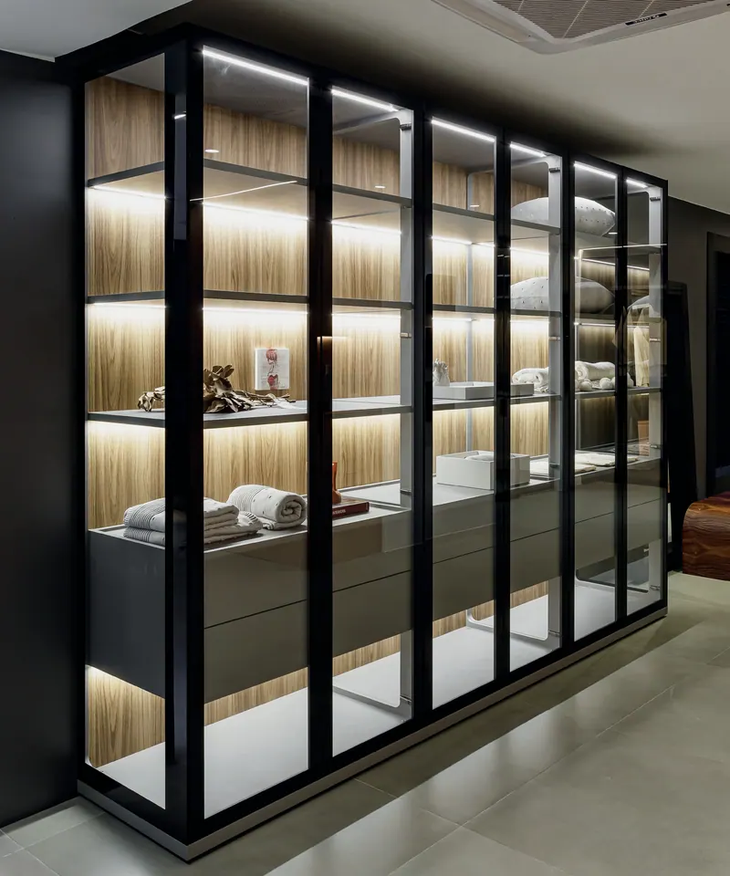 Armário de closet com portas de vidro e perfis pretos, prateleiras iluminadas e fundo em tom amadeirado