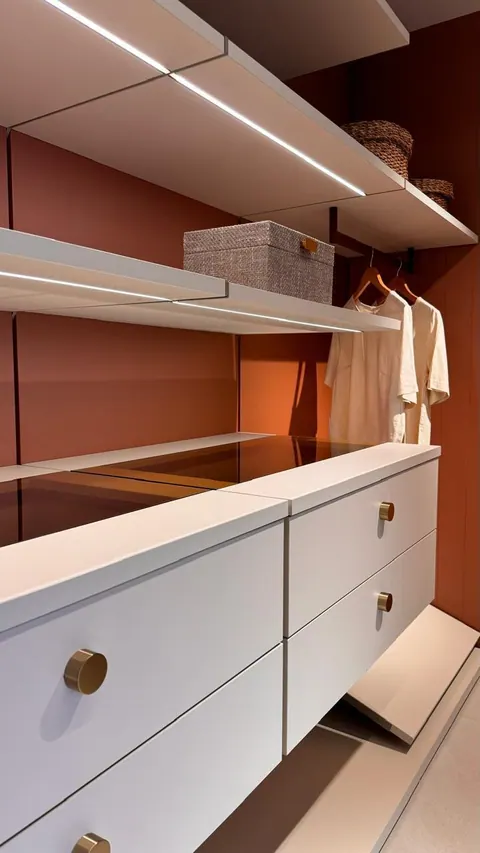 Closet aberto com prateleiras claras, fita de led, cabideiro e gaveteiro com puxadores dourados